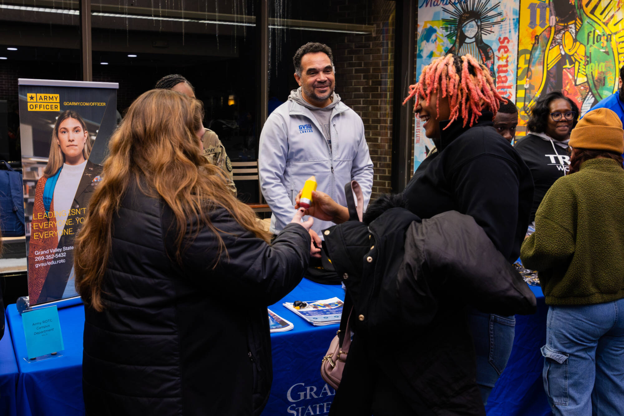 Two students smile at each other in front of a table where a man is smiling at them.